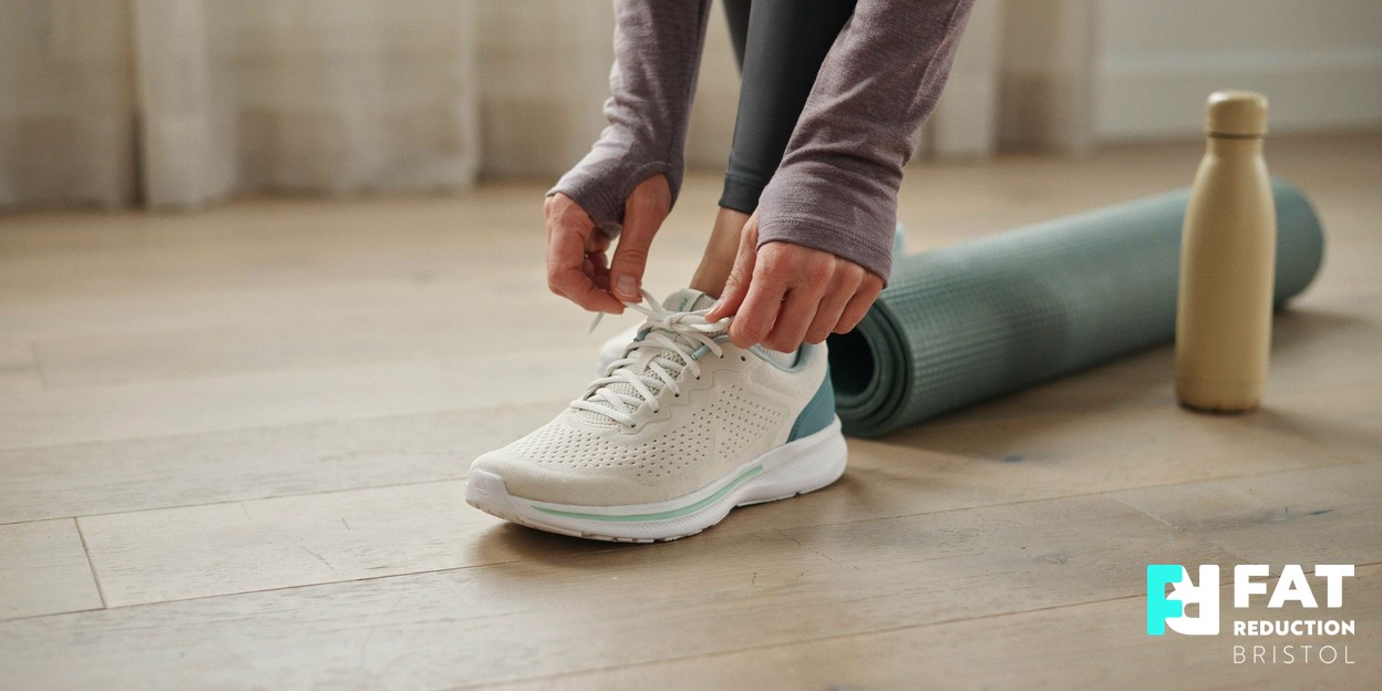 A person’s hands lacing up a clean athletic trainer on a light wooden floor, activewear and a rolled exercise mat softly blurred behind, an active-lifestyle moment