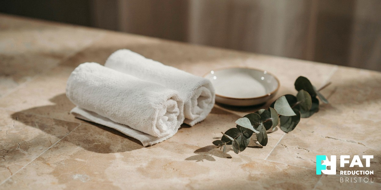 A soft-focus photograph of neatly rolled white towels and a small ceramic dish on a warm neutral spa surface in soft natural daylight