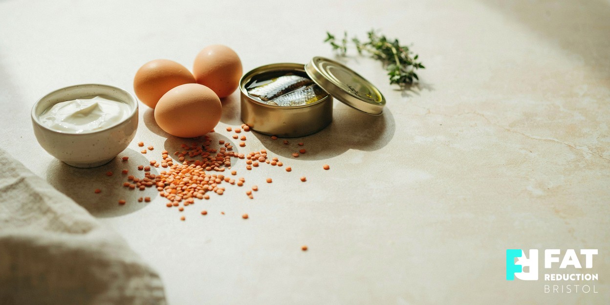 A photograph of a still-life of high-protein foods — eggs, a tin of fish and a bowl of Greek yoghurt — arranged on a light kitchen surface in soft daylight