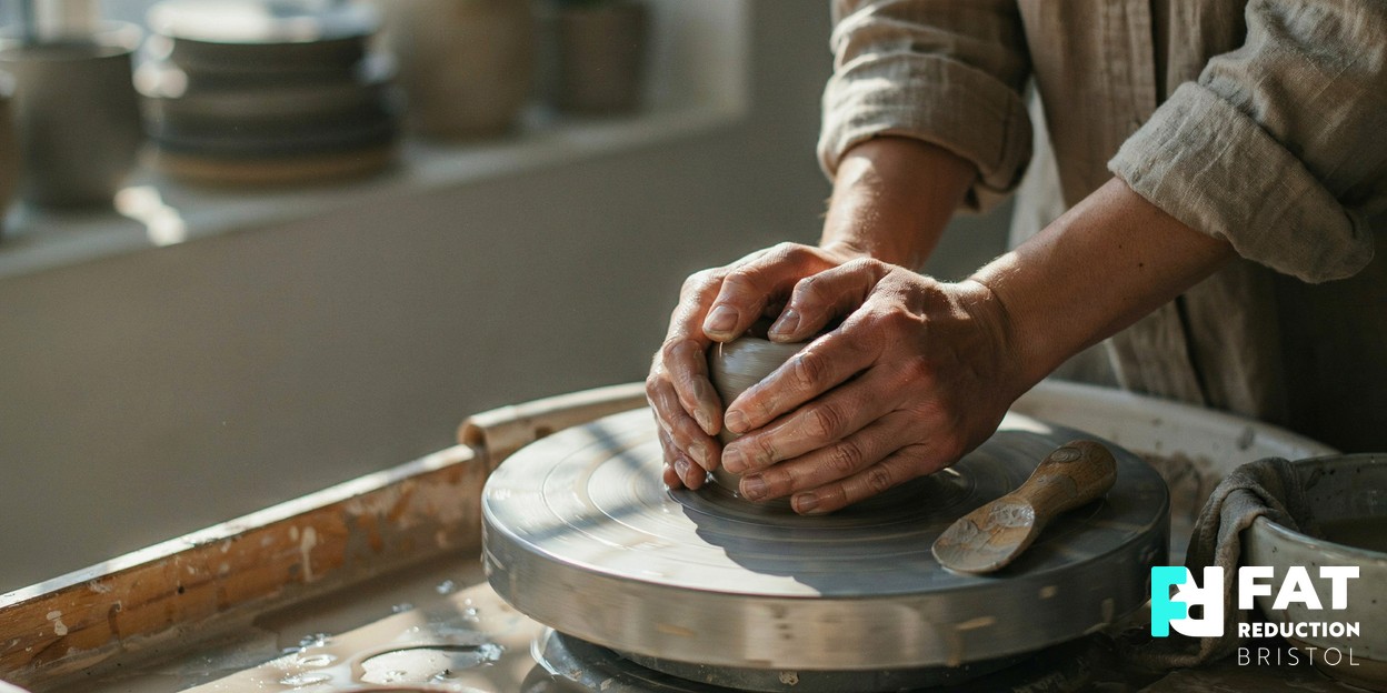 Photograph of a pair of hands gently shaping soft clay on a pottery wheel in warm daylight, symbolising careful body sculpting
